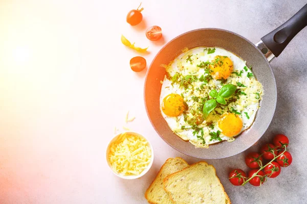 Sunny morning. Breakfast with fried eggs in pan, herbs, spices, cherry tomatoes, cheese toasts, basil on grey concrete stone background. Copy space, top view, flat lay, sunlights