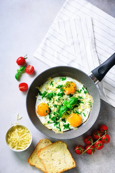 Fried eggs in pan, herbs, spices, cherry tomatoes, cheese toasts, basil for breakfast on grey concrete stone background. Copy space, top view, flat lay