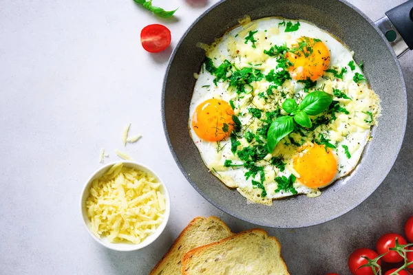 Fried eggs in pan, herbs, spices, cherry tomatoes, cheese toasts, basil for breakfast on grey concrete stone background. Copy space, top view, flat lay
