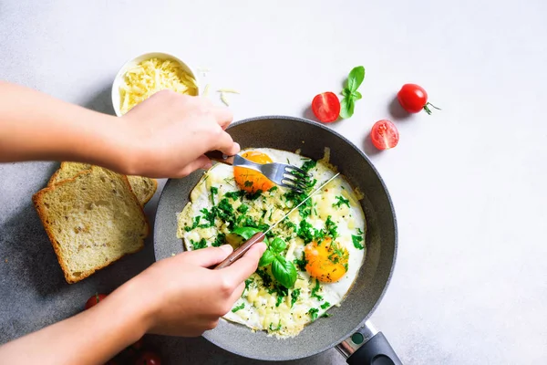 Girl hands above frying pan with three cooked eggs, herbs, cheese, tomatoes. Woman is making breakfast. top view. Copy space