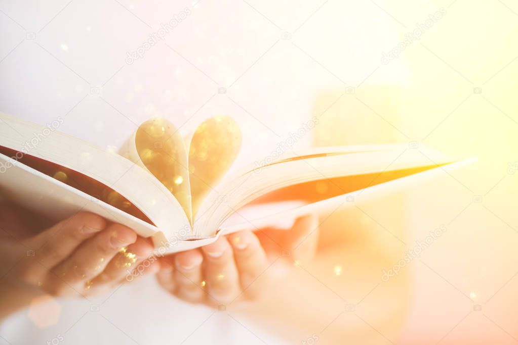 Book with opened pages and shape of heart in girl hands. Copy space. Love concept. Festive background with bokeh and sunlight. Magic fairy tale