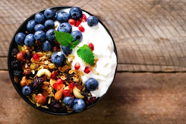 Tasty homemade granola, yogurt, fresh organic berries, pomegranate, mint on rustic wooden background with copy space, top view. — Stock Photo, Image