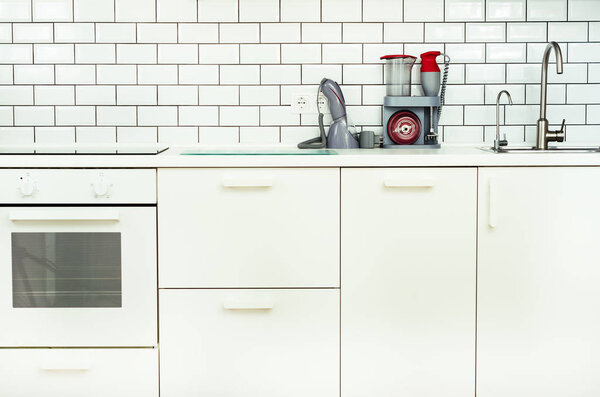 Белый минимализм интерьера и дизайна кухни. Плитка стены фона. Бытовая техника - блендер, вакуумная машина, современная плита, посудомоечная машина, духовка, стол, смеситель кранов

