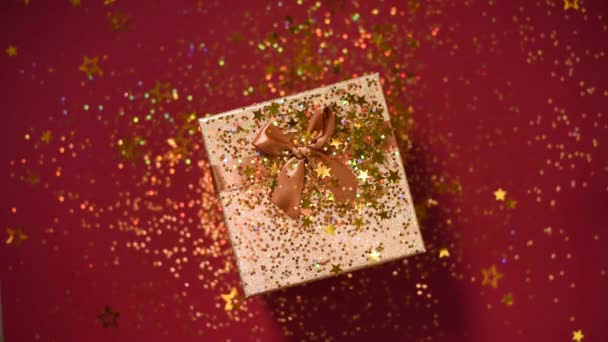 Sparkling gouden sterren, glitter confetti over geschenkdoos op rode achtergrond. Bovenaanzicht. Kerst en Nieuwjaar textuur. Verkoop, korting prijs, shopping concept. — Stockvideo