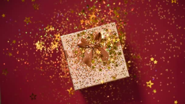 Sparkling gouden sterren, glitter confetti over geschenkdoos op rode achtergrond. Bovenaanzicht. Kerst en Nieuwjaar textuur. Verkoop, korting prijs, shopping concept. — Stockvideo