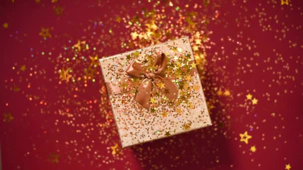 Sparkling gouden sterren, glitter confetti over geschenkdoos op rode achtergrond. Bovenaanzicht. Kerst en Nieuwjaar textuur. Verkoop, korting prijs, shopping concept. — Stockvideo