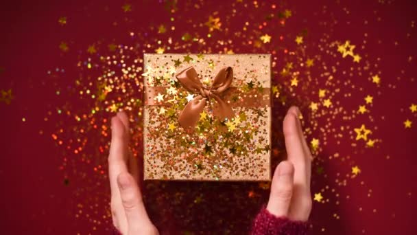 Kvinnliga händer öppnar presentförpackning. Mousserande guldstjärnor, glitter konfetti över närvarande. Packa upp gåva på röd bakgrund. Jul och ny årsmodell. — Stockvideo
