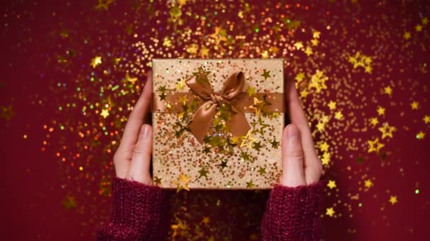 Les mains de femme ouvrant boîte cadeau. Étoiles d'or scintillantes, confettis scintillants sur le présent. Déballer cadeau sur fond rouge. Texture Noël et Nouvel An . — Video