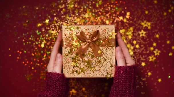 Kobiece dłonie otwierają pudełko. Błyszczące złote gwiazdki, błyszczące konfetti na prezent. Rozpakowywanie prezentów na czerwonym tle. Boże Narodzenie i Nowy Rok tekstury. — Wideo stockowe
