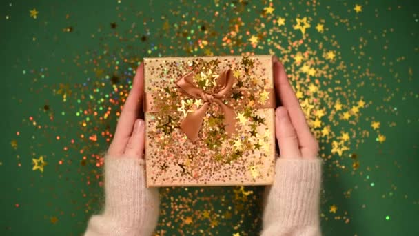 Ženské ruce otevírají dárkovou krabici. Jiskřivé zlaté hvězdy, třpytivé konfety v přítomnosti. Vybalování dárek na zeleném pozadí. Vánoční a novoroční textura.