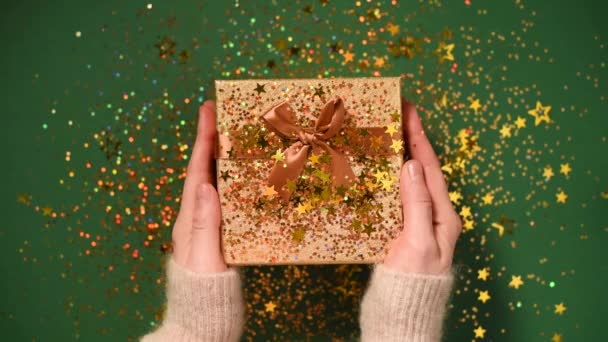 Woman hands opening gift box. Sparkling gold stars, glitter confetti over present. Unpacking gift on green background. Christmas and New year texture. — 비디오