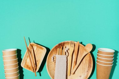 Bambu tabakları, tahta kaşık, çatal, bıçak, mavi arka planda kağıt bardaklar. Doğal malzemelerden tek kullanımlık sofra takımı. Sürdürülebilir yaşam tarzı konsepti. Çevre dostu tek kullanımlık aletler