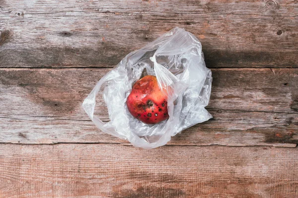 Spoiled bad red apple in plastic bag on wooden background. Garbage dump rotten food. Top view. Copy space. Rotten vegetables and fruits concept. Zero waste, compost