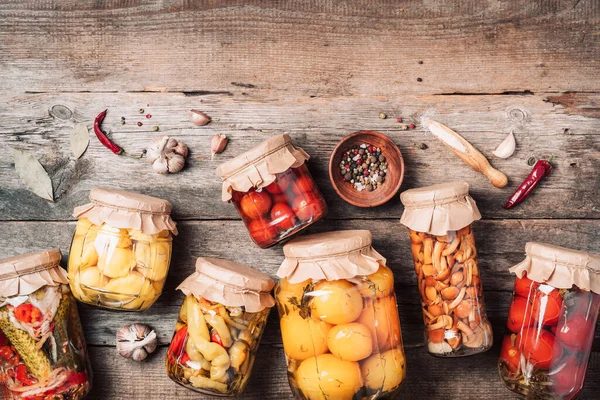 Canned and preserved vegetables in glass jars over wooden background. Top view. Flat lay. Copy space. — Stock Photo, Image