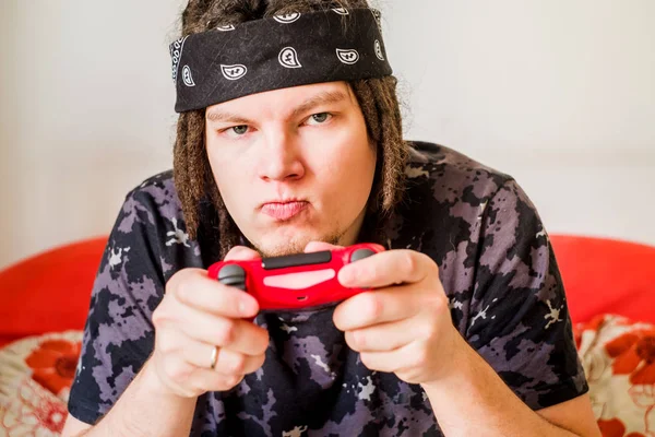 Junger Mann mit Dreadlocks und ernstem Gesicht, der Videospiele mit einem Steuerknüppel auf einer orangefarbenen Couch spielt — Stockfoto