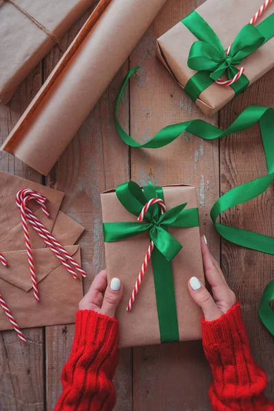 woman hands holding a christmas present wrapped with craft paper, green ribbon and candy cane top view on wooden table