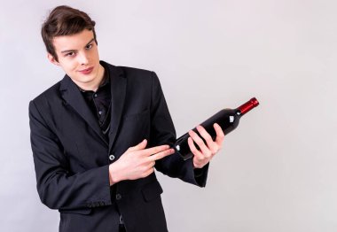 Genç adam şarap şişesini işaret ediyor, gri arka plandaki fotokopi çekilen kameraya bakıyor.