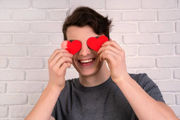 Молодой улыбающийся мужчина держит валентинки в форме сердца перед глазами на фоне белого кирпича — стоковое фото