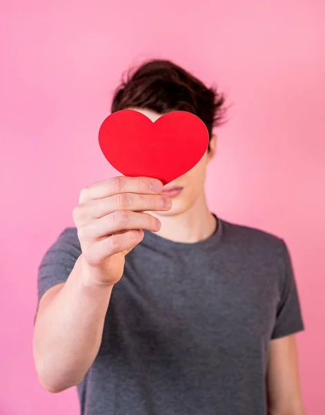 Молодой человек держит валентинку в форме сердца перед глазами на розовом фоне с копировальным пространством — стоковое фото