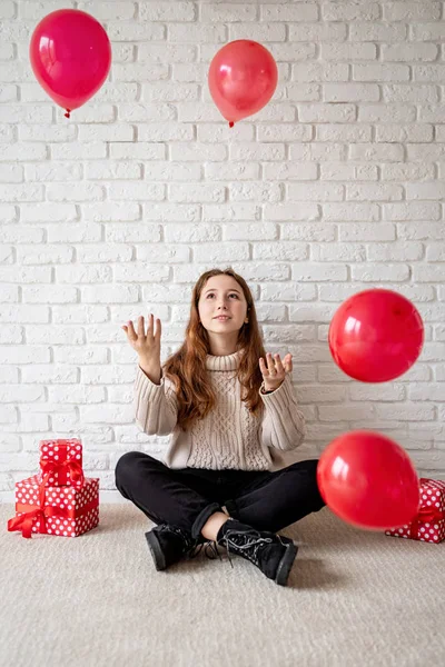 Schattige glimlachende jonge vrouw zittend op de beige tapijt gooien rode ballonnen in de lucht — Stockfoto