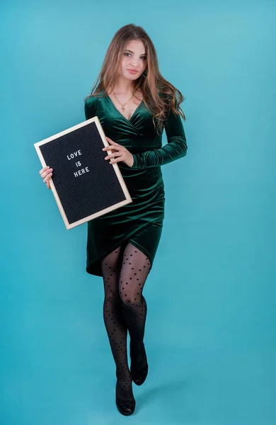 Vacker kvinna i grön klänning håller en filt brev styrelse med orden Kärlek är här på blå bakgrund — Stockfoto