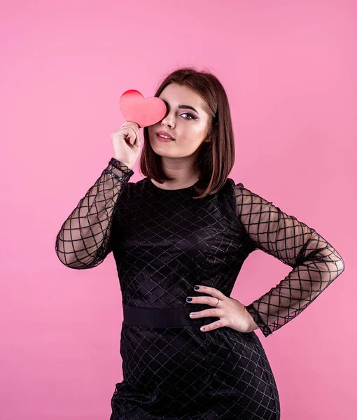 Sexy kobieta w czarny sukienka gospodarstwa serca kształt przed jej twarz izolowane na różowy tło — Zdjęcie stockowe