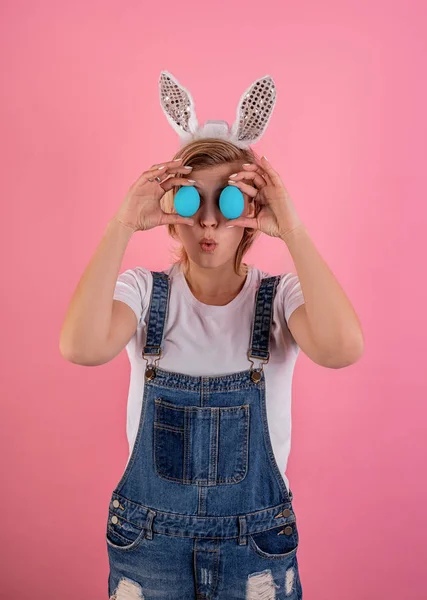 Mujer joven en orejas de conejo sosteniendo huevos de pollo de color frente a sus ojos aislados sobre fondo rosa — Foto de Stock