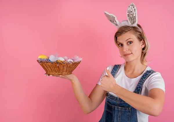 Junge Frau mit Hasenohren, die Daumen nach oben zeigt und einen Korb mit Ostereiern auf rosa Hintergrund hält — Stockfoto