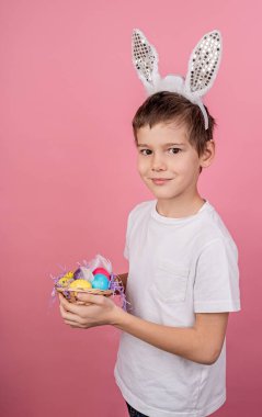 Smiling boy in bunny ears holding a basket with colorful eggs isolated on pink bakground