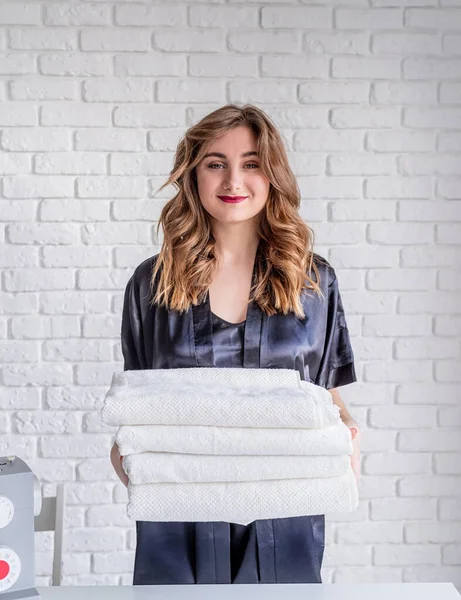 Beautiful young woman is holding a pile of clean white towels, looking at camera and smiling — Stockfoto