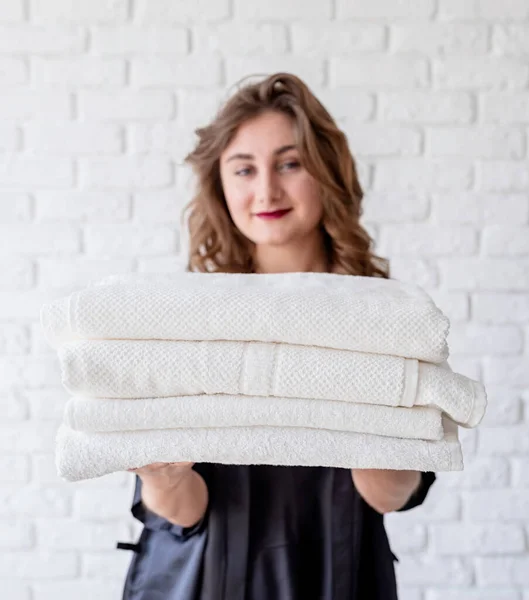 Smiling young woman holding a pile of towels on white bricks background — Stockfoto