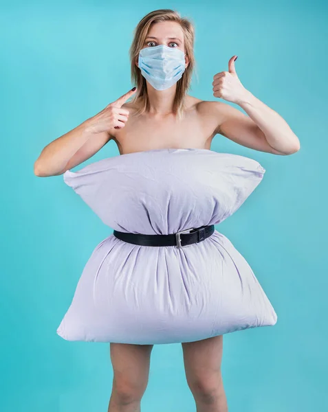 Coronavirus quarantine. Crazy quarantine. Blond woman in pillowdress wearing a mask showing thumbs up isolated on blue background. Pillow challenge outbreak