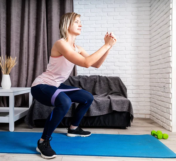 Stay home. Home fitness. Woman exercising squats with rubber resistance band at home