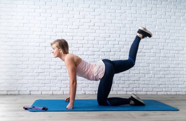 Evde kal. Evde fitness. Gülümseyen genç sporcu kadın dört ayak üstünde egzersiz yapıyor. Sırtüstü uzanıyor, bacağını içeride doğrultuyor.