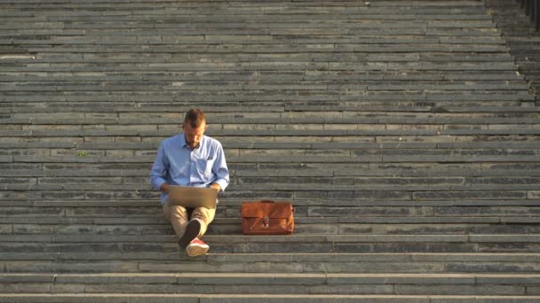 Vista lateral do jovem sentado nas escadas no parque usando laptop — Vídeo de Stock