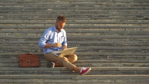 Ein junger Mann sitzt auf einer Treppe und arbeitet am Laptop. — Stockvideo