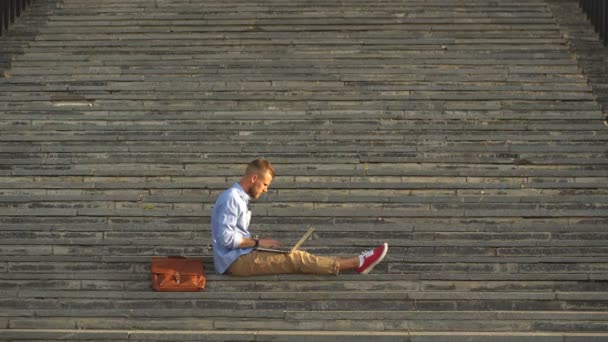 Красивий студент сидить на кам'яних сходах і використовує ноутбук — стокове відео