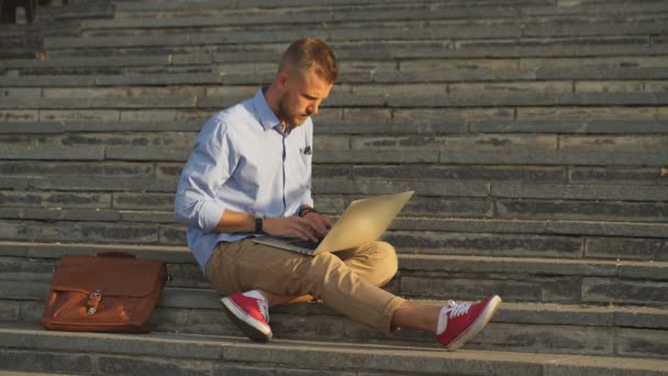 Estudiante guapo sentado en escalones de piedra y usando un portátil — Vídeo de stock