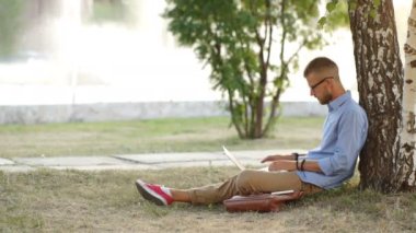 Genç sakallı adam bahçesinde ağaç altında yeşil çimenlerin üzerinde oturan ve onun laptop içinde çalışma