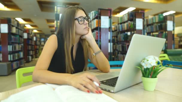 Student som uttråkad i biblioteket, arbetar på datorn i biblioteket, brunett med glasögon — Stockvideo