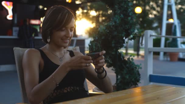 Beautiful woman sitting in a cafe at sunset and typing the message — Stock video