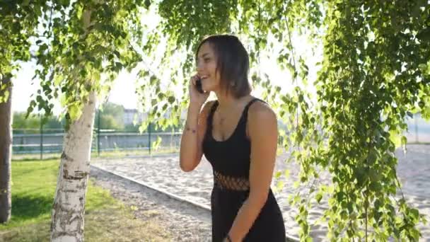 Cheerful brunette talking on the phone under a tree in a park — Stock Video