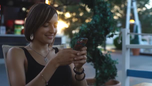 Красивая женщина сидит в кафе на закате и печатает сообщение — стоковое видео