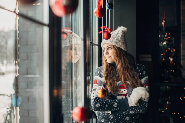 Beautiful brunette in Christmas sweater looking out the window — Stock Photo, Image