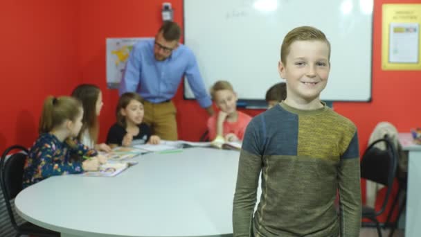 Little schoolBoy gesto polegares para cima enquanto segurando livros com colegas de classe estudando em segundo plano — Vídeo de Stock