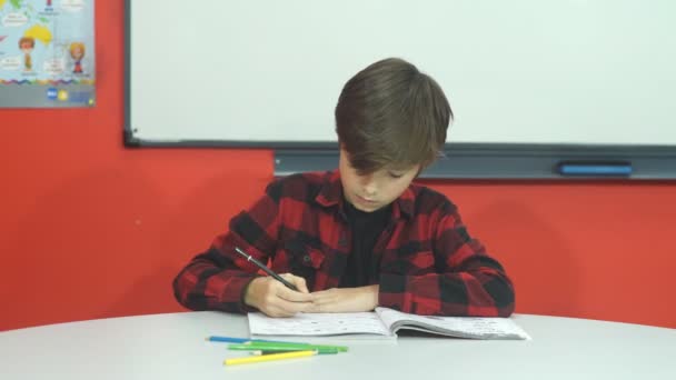 The student sits in class and writes in a notebook, looking out the frame — стоковое видео