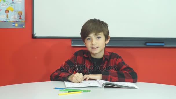 Menino da escola bonita está sentado na sala de aula e sorrindo para a câmera — Vídeo de Stock