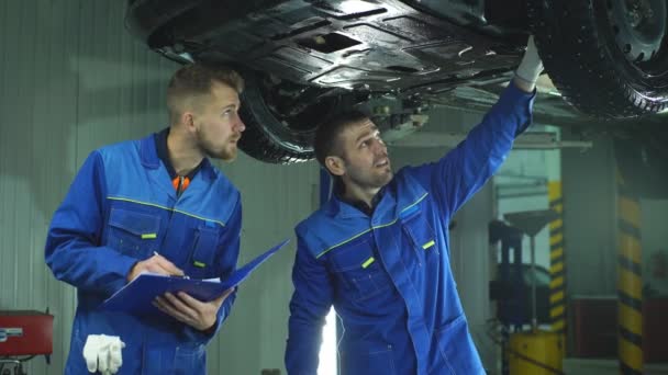 Молодые сотрудники мастерской работают вместе под поднятым автомобилем — стоковое видео
