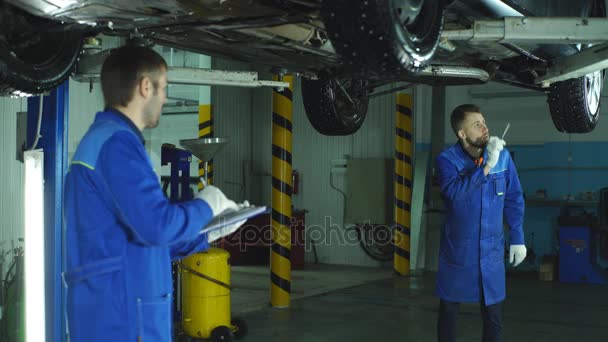 Два механика проверяют двигатель автомобиля и делают заметки — стоковое видео