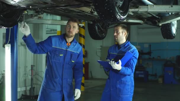 Mužské mechaniky pomocí digitálních tabletu při práci pod vyklopeným auto v auto opravna — Stock video